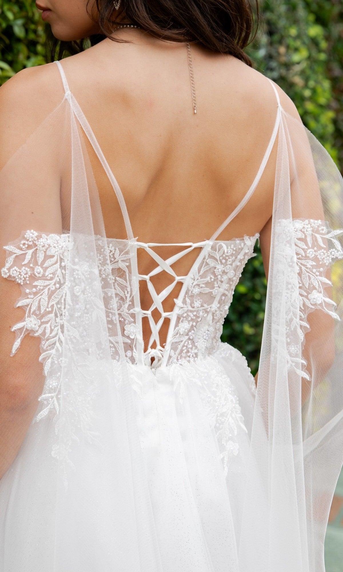A woman wears the Long Formal Dress: Cinderella Couture 4217, a white lace-up bridal gown featuring sheer off-shoulder sleeves, floral embroidery, a flowing tulle A-line skirt, and delicate pearl-beaded lace details, shown from the back.
